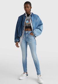 Chaqueta bomber de mezclilla en azul claro con puños de canalé y un forro interior a rayas, llevada sobre un top corto gráfico negro y combinada con unos jeans skinny azul claro y zapatillas blancas.