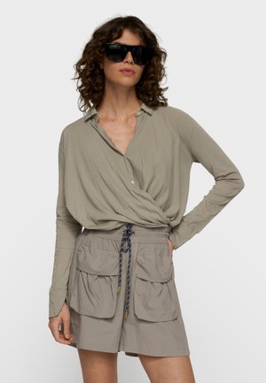 Mujer con pelo rizado que lleva grandes gafas de sol negras, camisa cruzada beige de manga larga y pantalones cortos beige con grandes bolsillos delanteros y cordón.