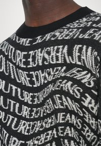 Zoom na črno pulover s bele popačene ponavljajoče se besedilo "JEANS COUTURE" okoli ovratnika in območja prsih.