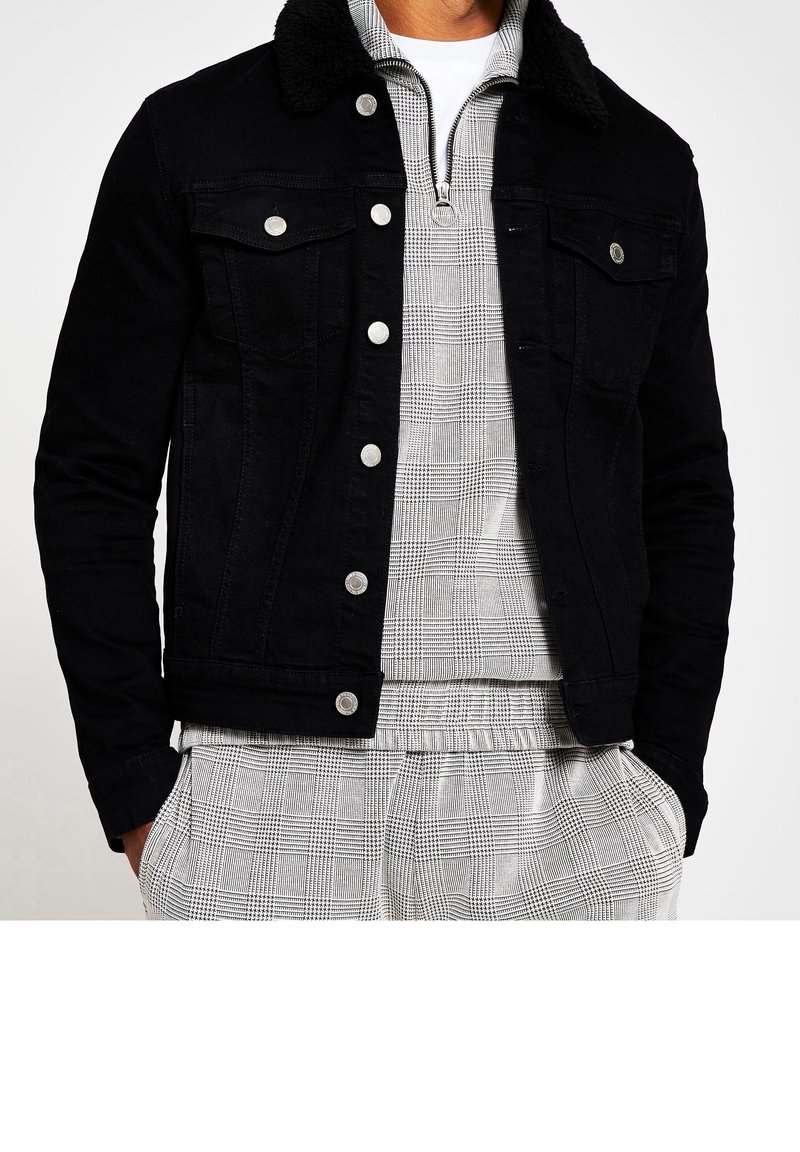 Man som bär en svart knappad jacka med fårskinnskrage över en grå och vit rutig zip-up-tröja och matchande byxor med händerna i fickorna.