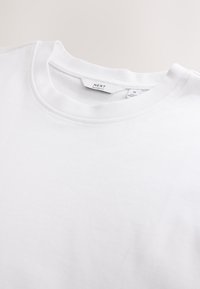 Weißes Baumwoll-T-Shirt mit Rundhalsausschnitt, mit "NEXT" Markenlabel und Größenetikett, das mittlere und lockere Passform-Details zeigt.