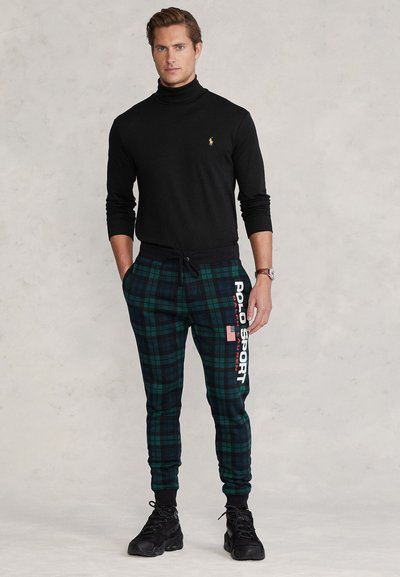 Mees, kes kannab musta kaelusega kampsunit ja rohelise ruudulise mustriga dressipükse valge "Polo Sport Ralph Lauren" tekstiga, seisab käed taskutes.