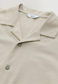 Chemise beige à boutons avec un col, en tissu texturé, deux boutons gris et une étiquette indiquant "NEXT REGULAR FIT".