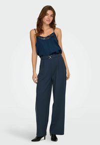 Marineblå satin camisole med palietter langs halsen, parret med højtaljede, brede bukser og spidse sorte ankelstøvler. Guld bæltet som accent.