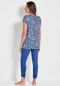 Vrouw staat blootsvoets met de rug naar ons toe, gekleed in een abstract blauw-grijs patroon top en blauwe gerimpelde enkelleggings met linten.