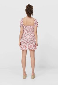 Kurzes Kleid mit Puffärmeln und quadratischem Ausschnitt, mit weißem Hintergrund und kleinen roten Blumenmustern. Rüschenunterkante und figurbetonte Silhouette.