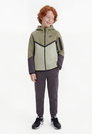 Rødharret dreng iført oliven- og grå Nike-hættetrøje, matchende joggingbukser og sort-hvide sneakers, stående med hænderne i lommerne.