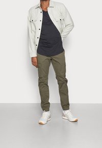 Uomo che indossa una giacca buttoned grigia chiara sopra una maglietta nera, pantaloni cargo color oliva e sneakers bianche, in piedi contro uno sfondo grigio.