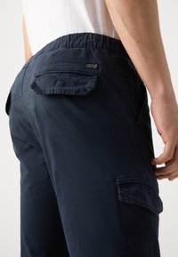 Homme portant un pantalon cargo bleu marine avec des poches à rabat et une ceinture élastique, debout devant un fond blanc uni.