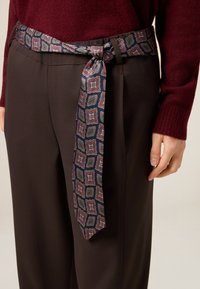 Jedwabny pasek w geometryczne wzory w kolorach granatowym, bordowym i zielonym, przewiązany wokół ciemnobrązowych spodni, dodający kontrastu wizualnego i tekstury.