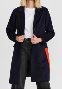 Abrigo de lana azul marino con corte entallado, que presenta un bolsillo de acento en naranja brillante, solapas tipo notch y dos botones frontales. Jeans de mezclilla gris por debajo.