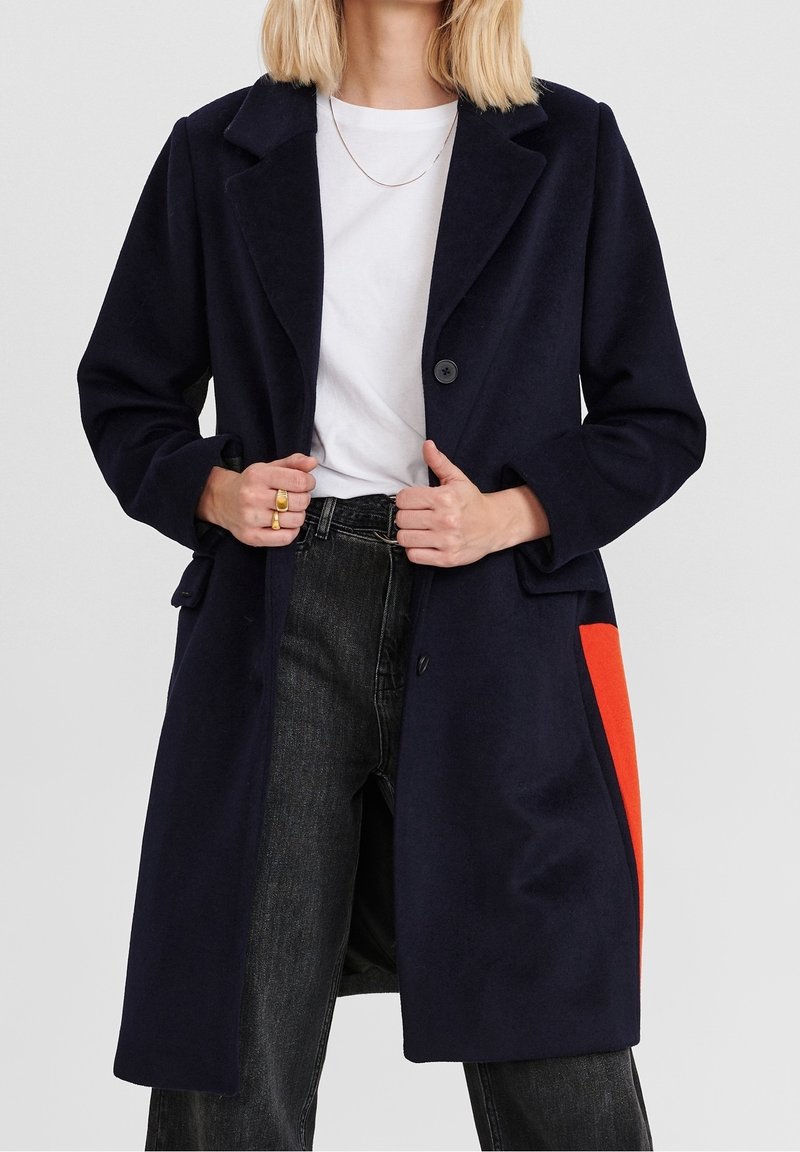 Abrigo de lana azul marino con corte entallado, que presenta un bolsillo de acento en naranja brillante, solapas tipo notch y dos botones frontales. Jeans de mezclilla gris por debajo.