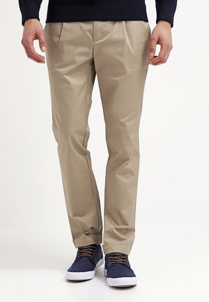 Mann trägt beige plissierte Hose, marineblauen Pullover und marineblaue Sneaker mit weißen Sohlen, steht vor schlichtem Hintergrund.