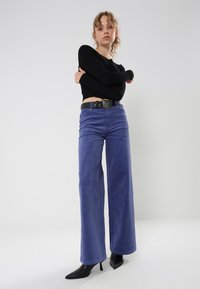 Pantalones de pana morados de pierna ancha con una cintura alta, bolsillos frontales y una distintiva hebilla plateada en forma de corazón en un cinturón negro.