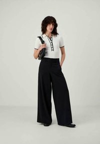Woman standing with one hand in pocket, wearing white short-sleeve collared top with black trim, black wide-leg pants, holding black shoulder bag.