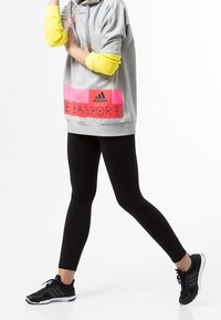 Sweat à capuche gris avec un panneau logo rose vif, superposé à un haut à manches longues jaune ; associé à des leggings noirs et des baskets noires.