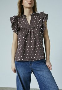 Modelen bærer en brun skjorte med prikker og flæser på ærmerne og kraven, som er sat sammen med løse blå jeans, mod en lys baggrund.