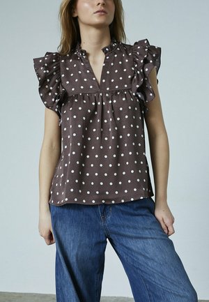 Modelen bærer en brun skjorte med prikker og flæser på ærmerne og kraven, som er sat sammen med løse blå jeans, mod en lys baggrund.