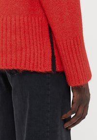 Maglione rosso lavorato a maglia con texture a coste, caratterizzato da un retro più lungo e fessure laterali. Abbinato a pantaloni grigio scuro, evidenziando il contrasto.