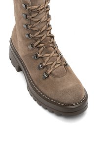 Bota de senderismo de ante marrón con suela gruesa y con tacos, y ojetes metálicos en forma de D, que presenta cordones redondos con un patrón atados en estilo entrelazado.