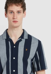 Korte mouwen shirt met afwisselende marineblauwe en lichtgrijze verticale strepen, met een button-down kraag en een klein oranje logo op de borst.