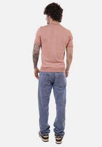 Uomo con il braccio destro tatuato che indossa una camicia rosa a maniche corte, jeans azzurri chiari e scarpe da ginnastica Nike marroni, visto di spalle.