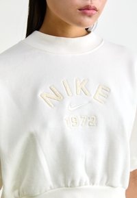 Sweatshirt branco com gola alta, apresentando bordados suaves em bege de "NIKE" e "1972", com cintura franzida para um ajuste perfeito.