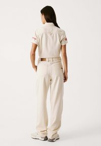 Camicia color crema a maniche corte con ricami rossi, abbinata a pantaloni beige a vestibilità rilassata. Presenta un design con colletto e gambe dritte.