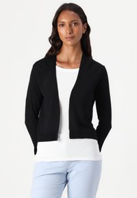 Zwarte gebreide cardigan met een sjawl-kraag, open voorkant en lange mouwen, gedragen over een witte geribde top, in combinatie met lichtblauwe broek.