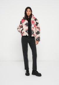 Flísová bunda v krémovej farbe s červenými a čiernymi geometrickými vzormi, zapínanie na zips a vysoký golier. Kombinovaná s čiernymi džínsami a robustnými čiernymi topánkami.