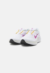 Zapatillas deportivas blancas con parte superior perforada, acentos morados y cordones amarillos. Suela de goma texturada negra para agarre y estabilidad.
