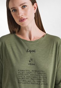 Grünes, leichtes, kurzärmliges Hemd mit rundem Ausschnitt. Mit gedrucktem Text in Schwarz, einschließlich "Repeat" und dekorativen Stilwörtern.