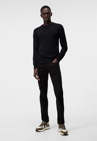 Juodas megztinis megztas, derinamas su juodomis kelnėmis, turinčiomis liekną siluetą. Modelis dėvi daugiomis spalvomis margintus sportinius batelius su stambiu dizainu ir tekstūruotais akcentais.