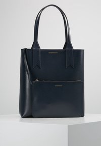 Marineblaue Ledertasche mit kontrastierender glatter Textur, ausgestattet mit zwei Griffen und einem abnehmbaren Reißverschlusstäschchen. Goldene Logodetails sichtbar.