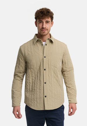 Man met kort krullend haar die een beige doorgestikte blouse met knopen draagt en een donkere broek, staand tegen een witte achtergrond.