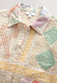 Besticktes Hemd mit pastellfarbenen geometrischen Mustern, cremefarbenem Hintergrund, Knopfleiste und einem Kragen. Der Stoff wirkt strukturiert.