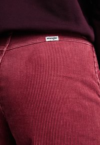 Pantaloni din corduroy burgundy cu bande verticale; au un eticheta albă "Wrangler" pe talie, evidențiind suprafața texturată și culoarea.