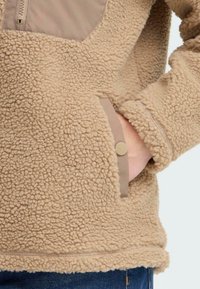 Veste en polaire beige avec un tissu texturé, des accents beiges, un zip half et un bouton-pression sur la manche. Comprend une poche kangourou.