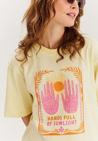 Žluté tričko s krátkým rukávem s růžovým a oranžovým grafickým designem dvou rukou a slunce, s dekorativními květinovými akcenty a textem.