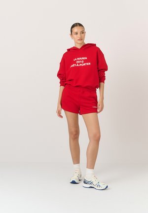 Czerwony hoodie z białym tekstem i pasującymi czerwonymi szortami. Materiał bawełniany, luźny krój, z kieszenią kangurzą w hoodie. Białe buty sportowe.