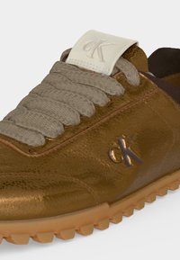 Baskets en cuir marron avec lacets beige texturés, étiquette blanche sur la languette et logo métallique "cK" sur le côté, avec une semelle en gomme robuste.