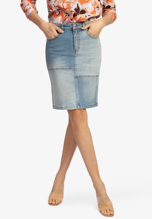 Frau trägt einen knieumspielenden, zweifarbigen Jeansrock mit Fronttaschen, kombiniert mit einer geblümten Bluse und klaren, offenen Zehenabsätzen.