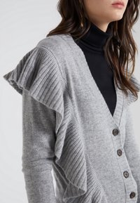 Cárdigan gris con textura acanalada y detalles de volantes en las mangas. Presenta un escote en V profundo y botones marrones en la parte frontal.