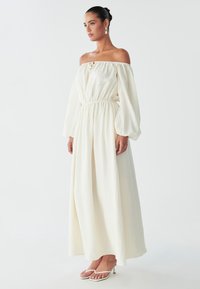 Una donna è in profilo, indossa un abito bianco fluente con scollatura off-the-shoulder e sandali bianchi con tacco, il tutto su uno sfondo minimalista.