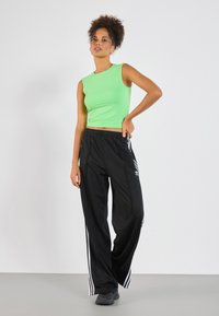 Green sleeveless crop top paired with black track pants featuring white side stripes and an Adidas logo. Smooth fabric, relaxed fit.