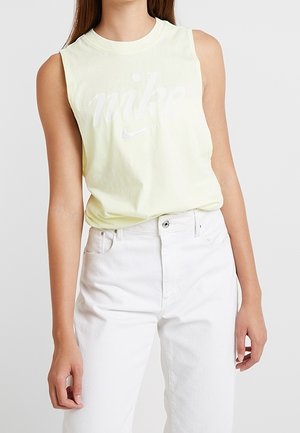 Bezrekawowy jasnółóżowy top Nike z białym logo w połączeniu z wysokim stanem białych jeansów noszonych przez stojącą osobę.