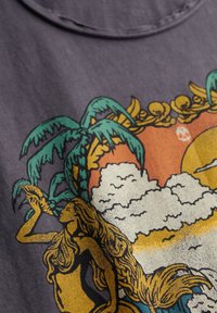 T-shirt gris avec un imprimé graphique coloré représentant une femme aux longs cheveux, entourée de palmiers et d'un soleil avec des nuages.
