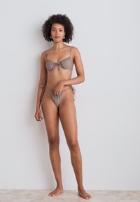 Rudas bikinio komplektas su surinktu viršumi ir kaspinu, reguliuojamais raiščiais, derinamas su aukštai iškirptomis kelnaitėmis su šoniniais raiščiais, pateiktas prieš neutralią fono spalvą.