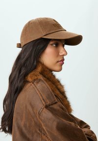 Cappello marrone con visiera curva e testo in rilievo, abbinato a una giacca marrone con colletto in pelliccia sintetica. Vista di profilo.