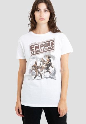 Frau mit mittelbraunen Haaren trägt ein weißes Star Wars The Empire Strikes Back T-Shirt mit Charakter- und Kreaturengrafiken auf der Vorderseite.